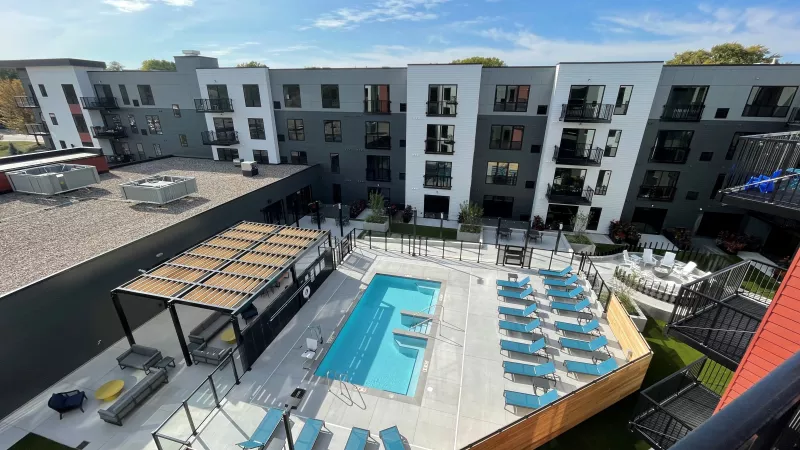 Modern apartment complex with a rectangular pool, light blue lounge chairs, and an outdoor bar on a sunny day.