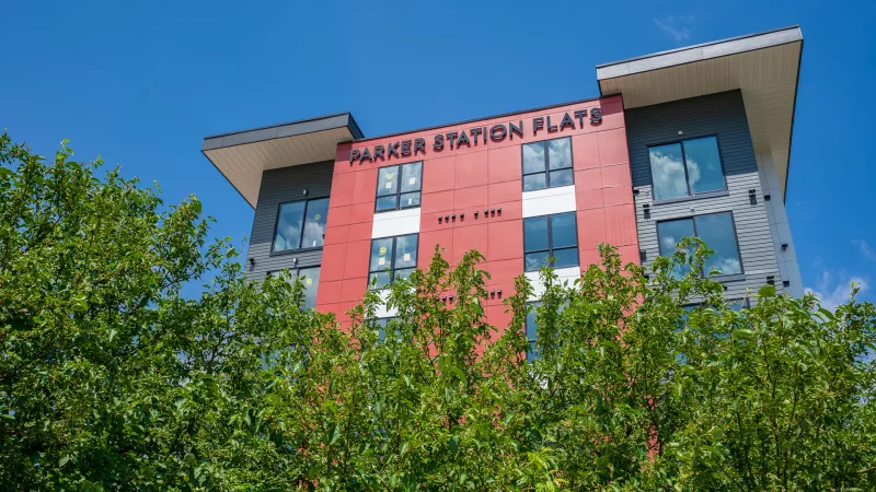 Modern multi-story building, "PARKER STATION FLATS" sign prominently displayed. Red, gray, and white facade, with green trees in front.