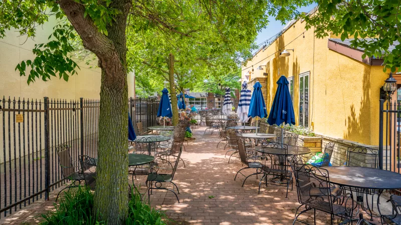 Sunny outdoor restaurant patio with empty metal tables, blue umbrellas, and a yellow building shaded by trees.