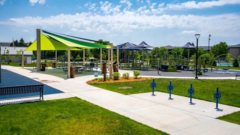 Sunny park with a large green shade canopy, blue umbrella structures, paved paths, and outdoor exercise equipment.