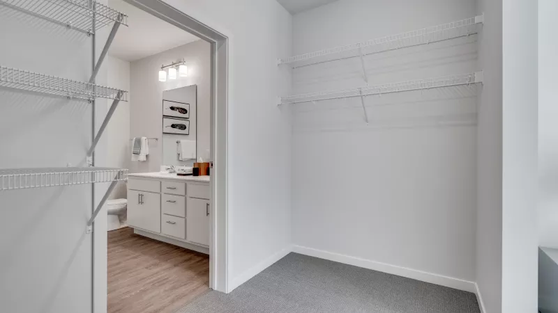 Empty walk-in closet with wire shelves, gray carpet, and white walls. An open doorway shows a bathroom vanity.