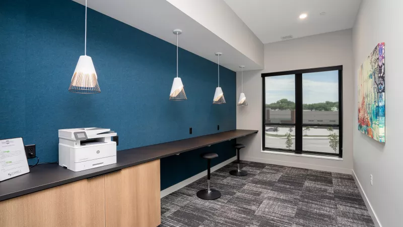 Modern study room with a long dark desk, blue wall, pendant lights, a printer, and a large window overlooking buildings.