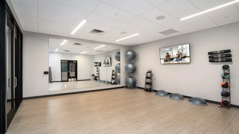 Bright, modern fitness room with a large mirror, stability balls, Bosu balls, and wall-mounted TVs showing workout videos.