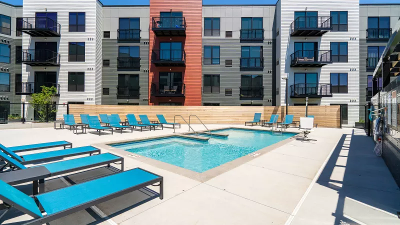 Modern apartment building with a sunny outdoor pool, hot tub, and teal lounge chairs on a concrete deck.