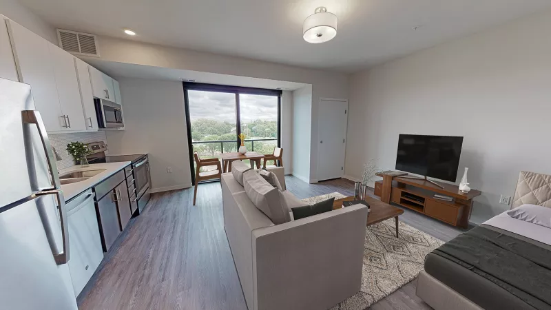 Modern furnished studio apartment with kitchen, living area, dining table, and bed. Large window overlooks green trees.