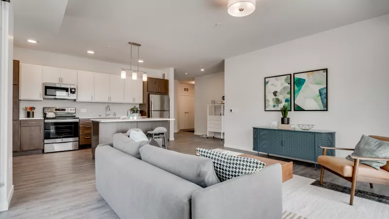 Modern open-plan living room and kitchen. Grey sofa, coffee table, teal console with art, and kitchen island with light cabinets.