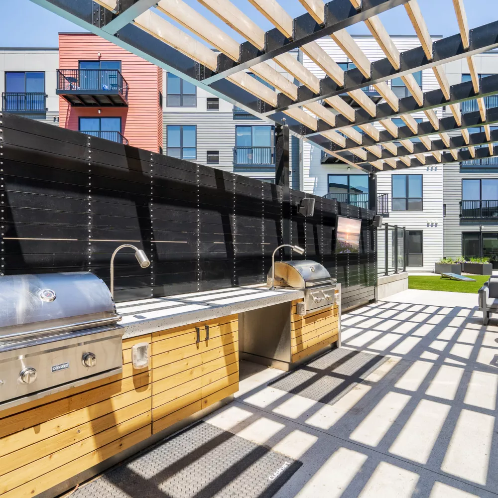 Photograph: Modern outdoor BBQ area with stainless steel grills, a sink, and wooden cabinets, shaded by a slatted pergola.