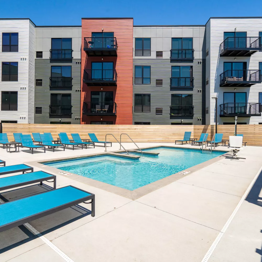 Modern apartment building with a sunny outdoor pool, hot tub, and teal lounge chairs on a concrete deck.