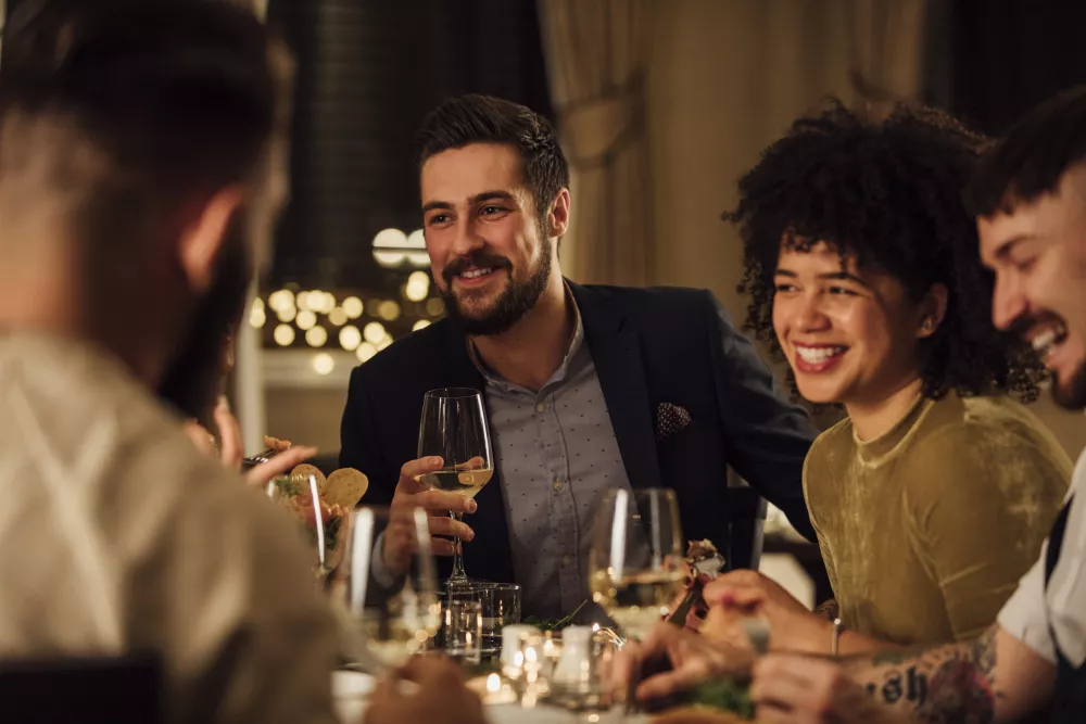Smiling friends enjoy white wine and conversation at a dimly lit dinner party, laughing happily.