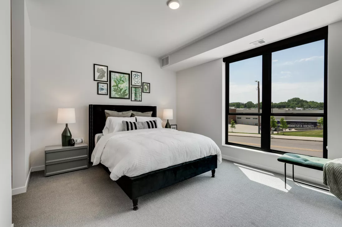 Bright modern bedroom with a dark bed, white duvet, green botanical art, and a large window overlooking a street.
